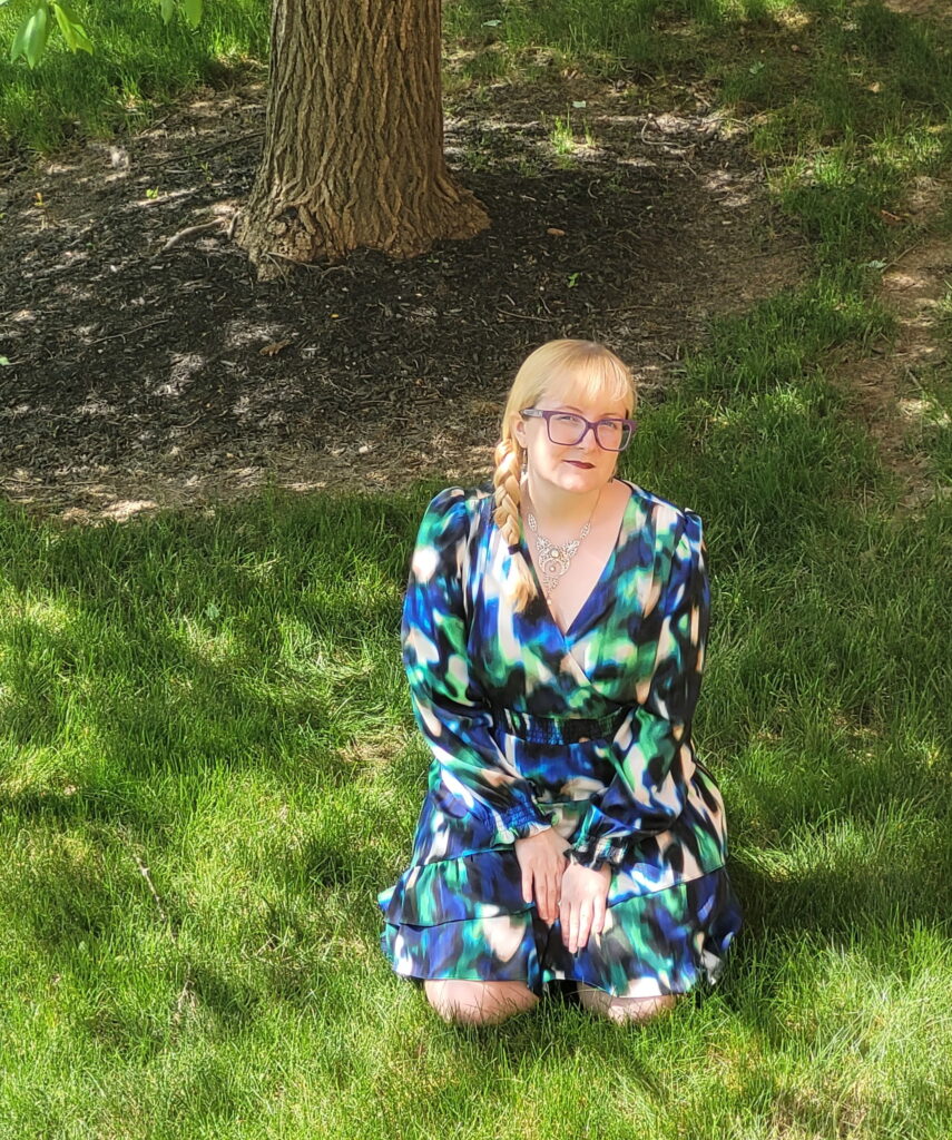 Photo of the author, Ashley Hawley, in a dress with blue, green, black, and white designs, leaning in the grass.