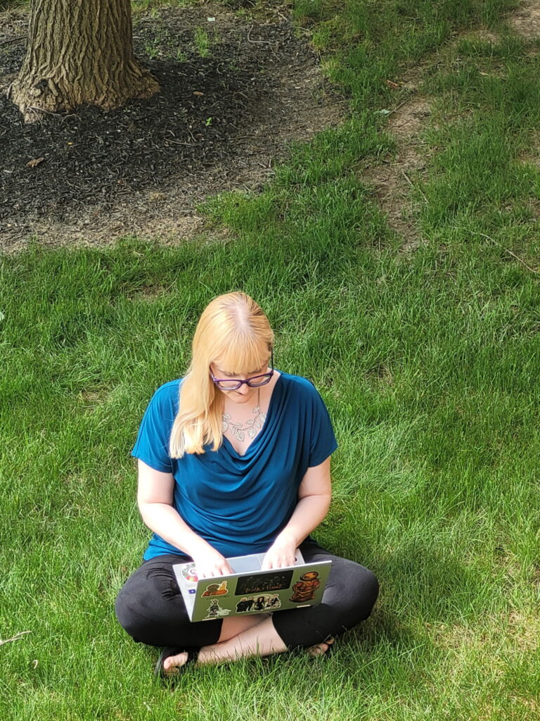 Photo of the author, Ashley Hawley, sitting in the grass on her laptop. She is wearing a dark teal blouse and black capris.